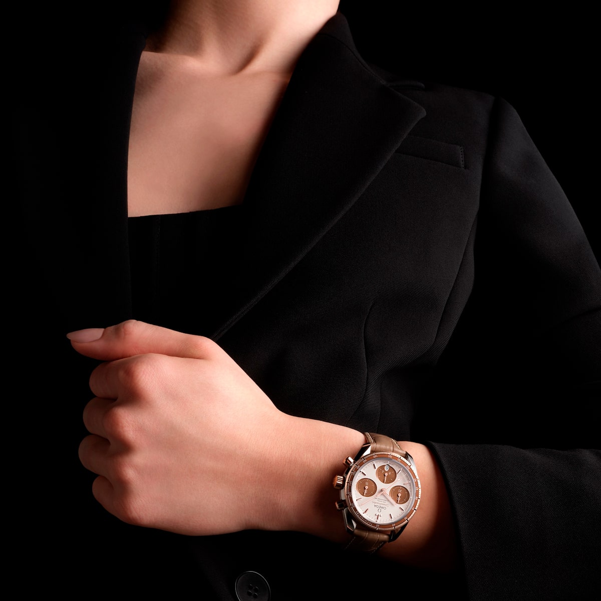 2025 © OMEGA Eine Person, die einen schwarzen Blazer trägt, hält mit einer Hand das Revers und zeigt eine elegante Speedmaster 38-Armbanduhr mit braunem Lederarmband und weißem Zifferblatt vor schwarzem Hintergrund.