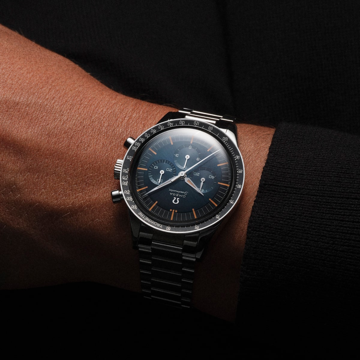 2025 © OMEGA Eine Nahaufnahme des Handgelenks einer Person, die eine Omega Speedmaster Heritage Uhr aus Edelstahl mit schwarzem Zifferblatt, Chronographenzifferblättern und einer Tachymeterlünette vor einem dunklen Hintergrund trägt.