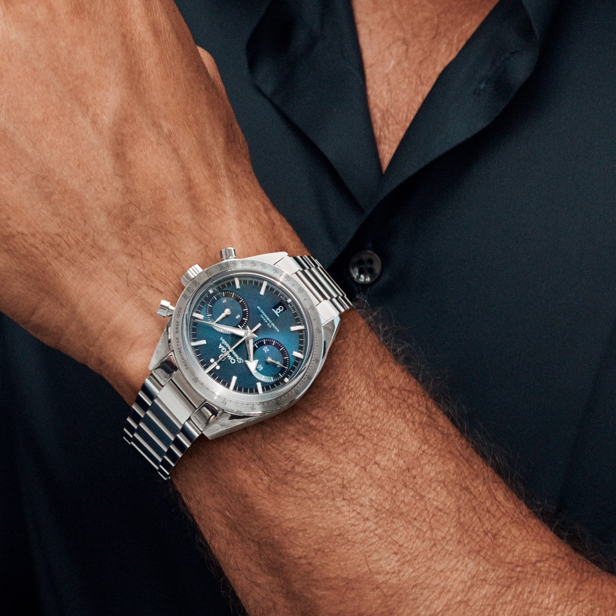 2025 © OMEGA Eine Person in einem dunklen Hemd trägt eine silberne Speedmaster Heritage Armbanduhr mit blauem Zifferblatt und mehreren Unterzifferblättern an ihrem linken Handgelenk. Die Uhr hat ein Metallarmband und Chronographenknöpfe.