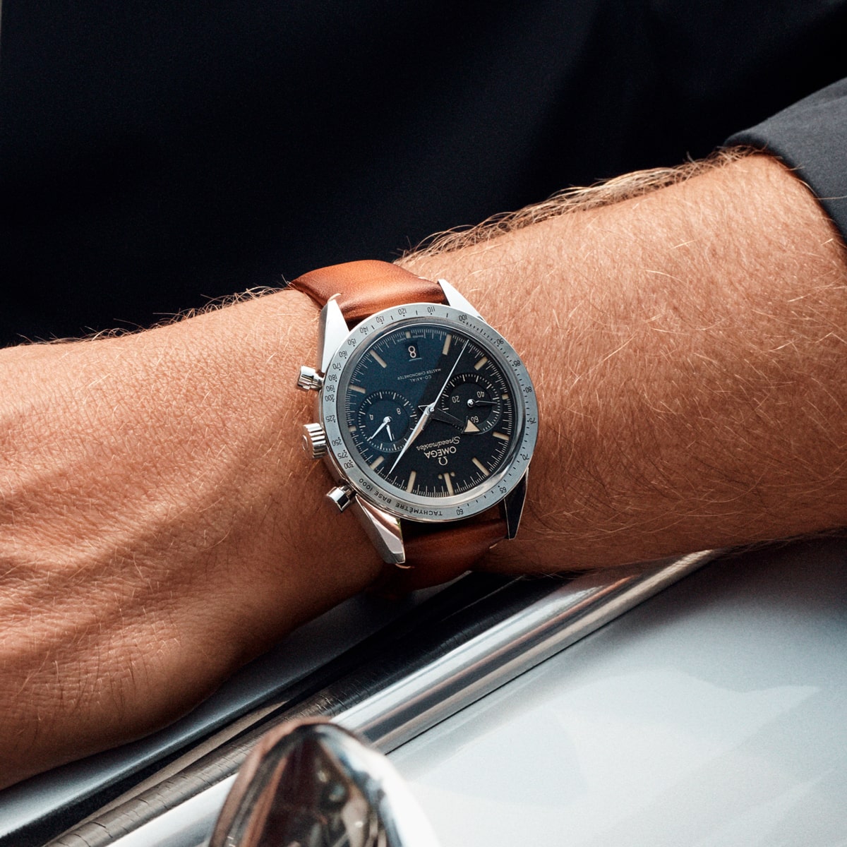 2025 © OMEGA Nahaufnahme des Handgelenks einer Person, die eine silberne Speedmaster Heritage Chronographenuhr mit blauem Zifferblatt und braunem Lederarmband trägt, die auf einer glänzenden Metalloberfläche liegt. Die Person ist mit einem dunklen Hemd bekleidet.