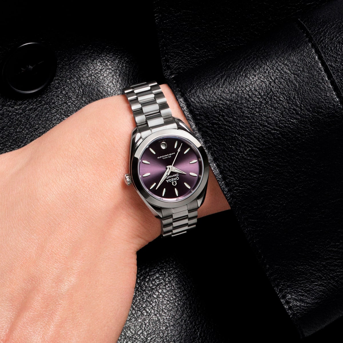 2025 © OMEGA Eine Person trägt eine silberne Aqua Terra Armbanduhr mit einem Metallarmband und einem dunkelvioletten Zifferblatt mit silbernen Stundenmarkierungen auf einem schwarzen Lederjackenärmel.
