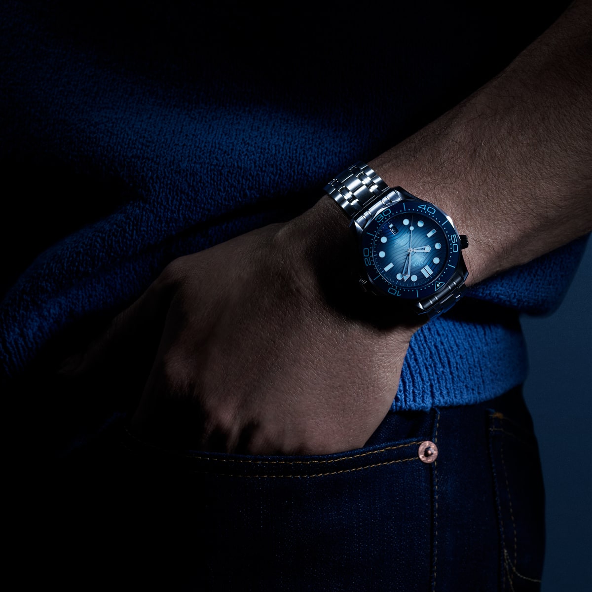 2025 © OMEGA Eine Nahaufnahme des Handgelenks einer Person, die eine silberne Diver 300M-Taucheruhr mit blauem Zifferblatt trägt, die teilweise beleuchtet ist. Die Person ist mit einem blauen Pullover und dunklen Jeans bekleidet und hat die Hand in die Tasche gesteckt.