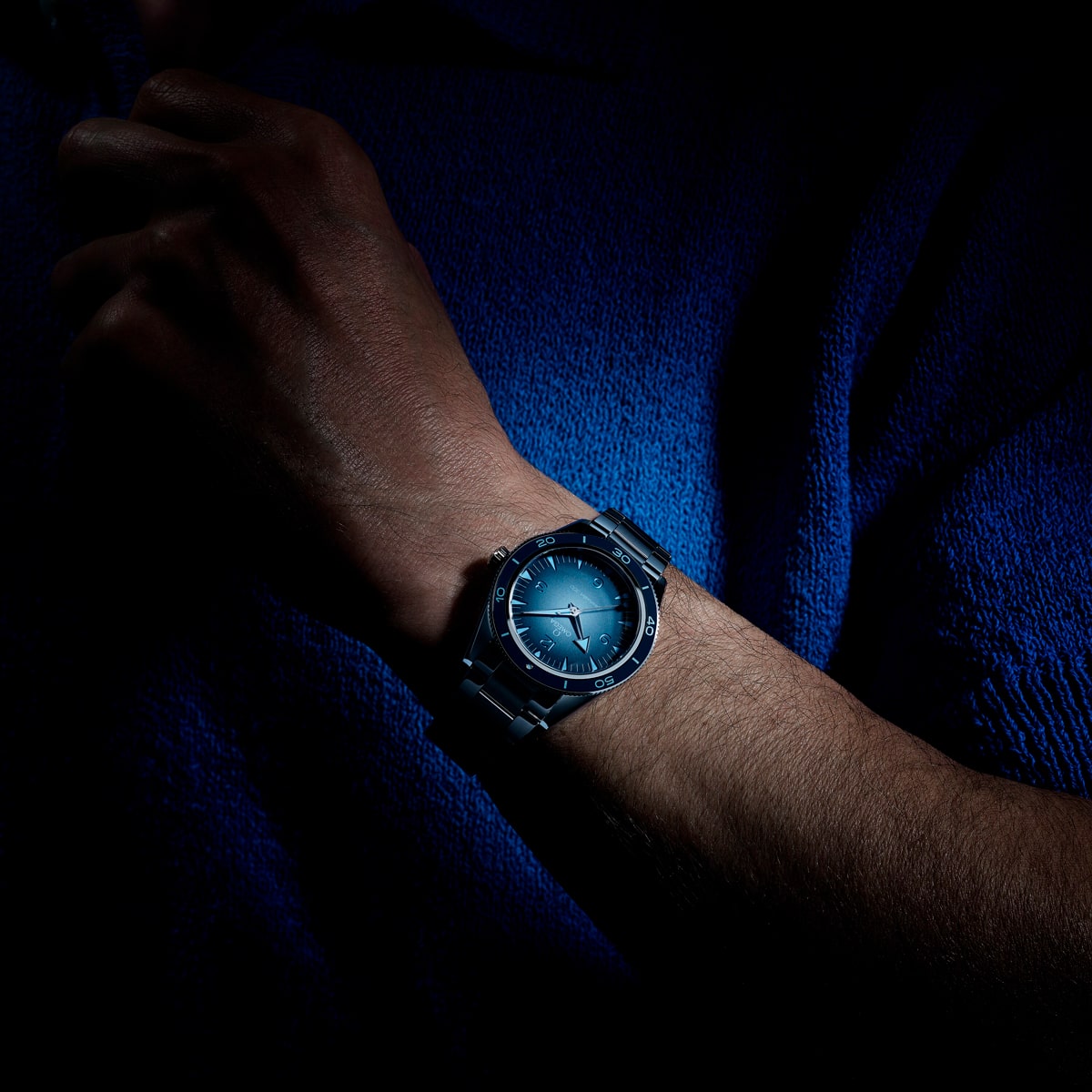 2025 © OMEGA Eine Nahaufnahme des Handgelenks einer Person, die eine silberne Seamaster Heritage-Armbanduhr mit blauem Zifferblatt trägt, vor einem dunklen Hintergrund mit dramatischer Beleuchtung, die die Uhr und einen Teil des Arms hervorhebt.