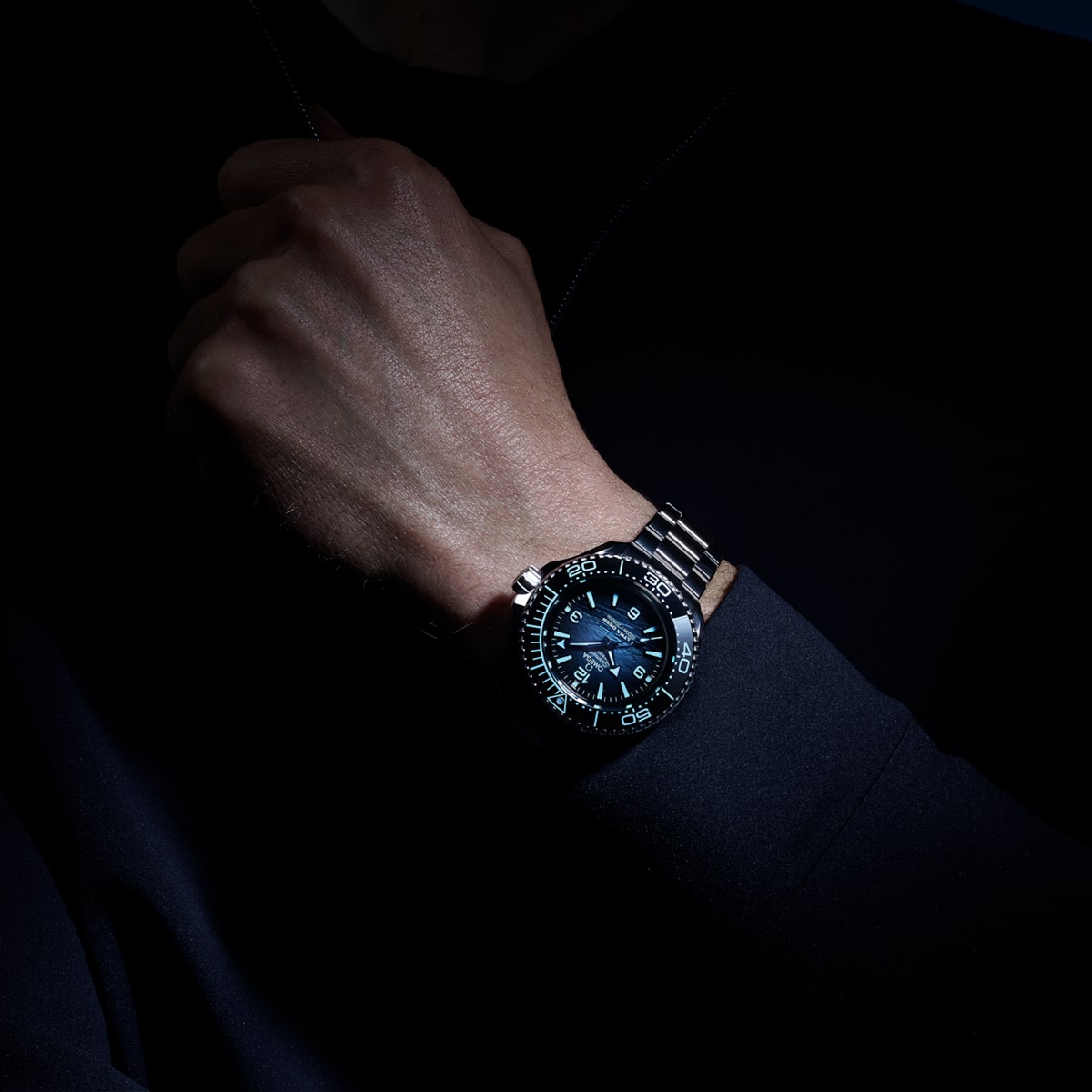 2025 © OMEGA Die linke Hand und das Handgelenk einer Person, die eine große, schwarze Planet Ocean Analoguhr mit einem Metallband trägt, sind vor einem dunklen Hintergrund hervorgehoben; Details des Ziffernblatts und der Lünette sind deutlich zu erkennen.