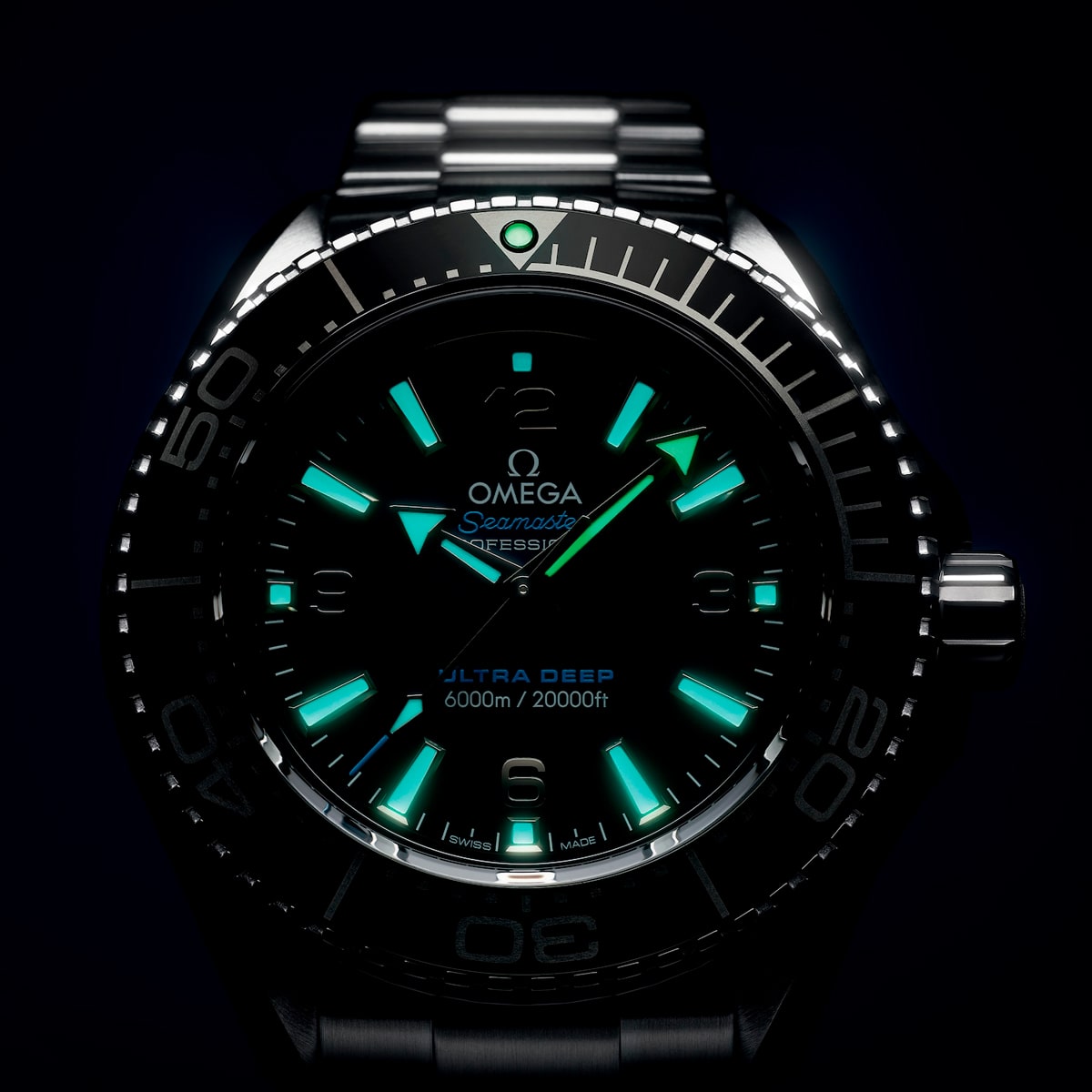 2025 © OMEGA Eine Nahaufnahme einer Omega Seamaster Planet Ocean Ultra Deep mit leuchtenden blauen und grünen Indexen und Zeigern, die die Tiefenangabe von 6000m/20000ft vor einem dunklen Hintergrund anzeigen.
