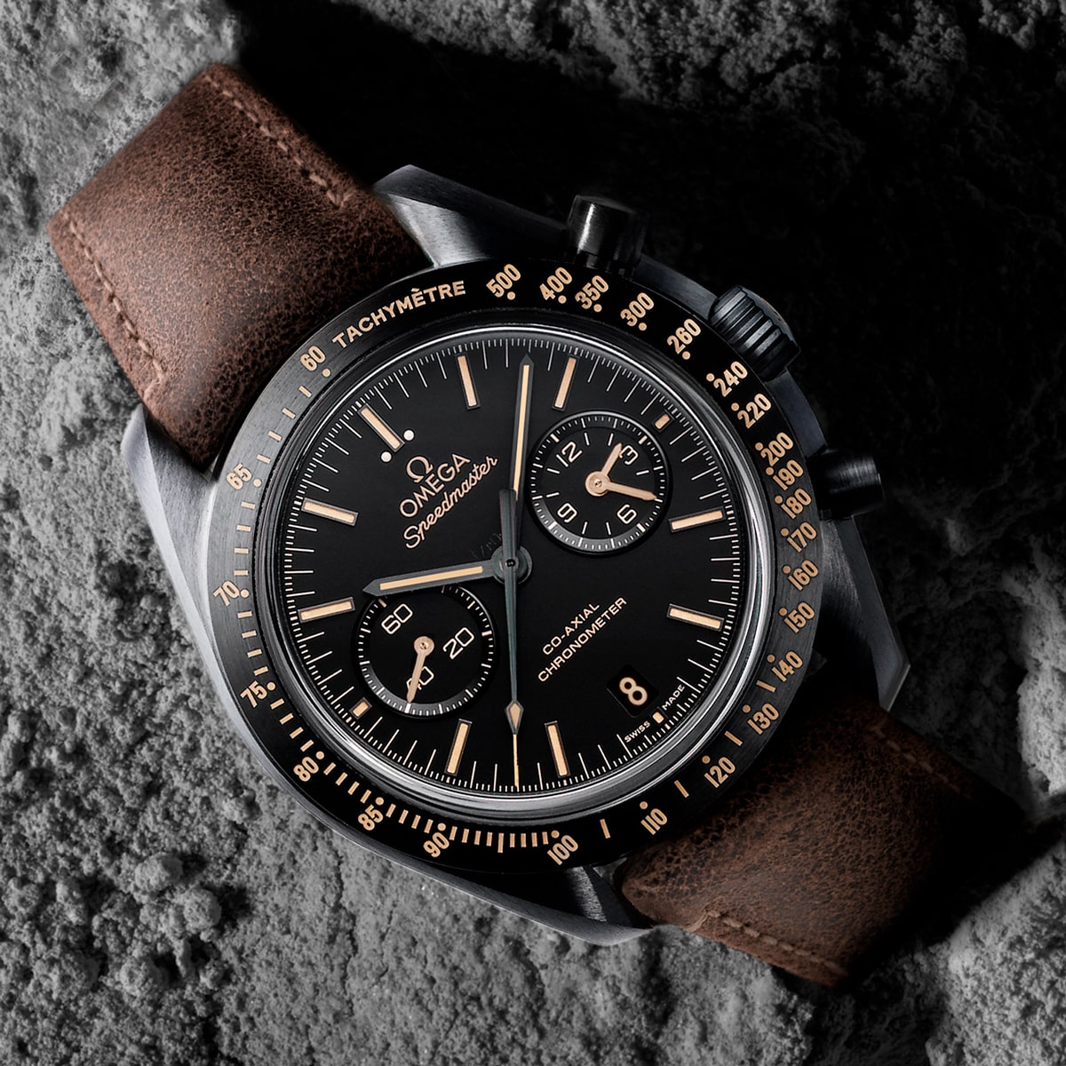 2025 © OMEGA Eine Nahaufnahme einer Omega Speedmaster Dark Side of the Moon Armbanduhr mit schwarzem Zifferblatt, goldenen Akzenten, braunem Lederarmband und Chronographenfunktionen auf einer strukturierten grauen Oberfläche, die an Mondgestein erinnert.
