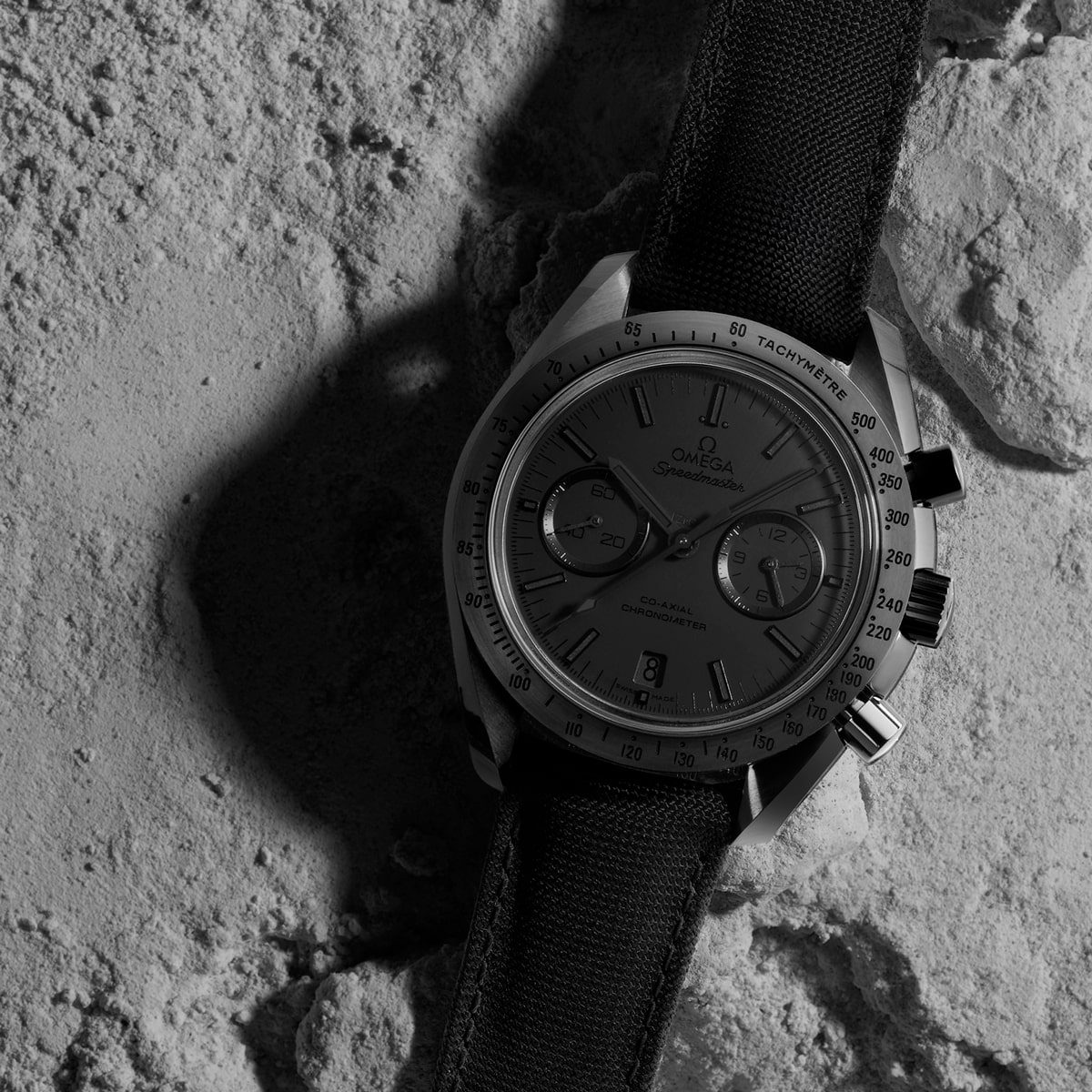 2025 © OMEGA Ein Schwarz-Weiß-Foto einer Omega Speedmaster Dark Side of the Moon-Armbanduhr mit Stoffarmband auf einer felsigen, mondähnlichen Oberfläche, wobei die Schatten die Details der Uhr hervorheben.