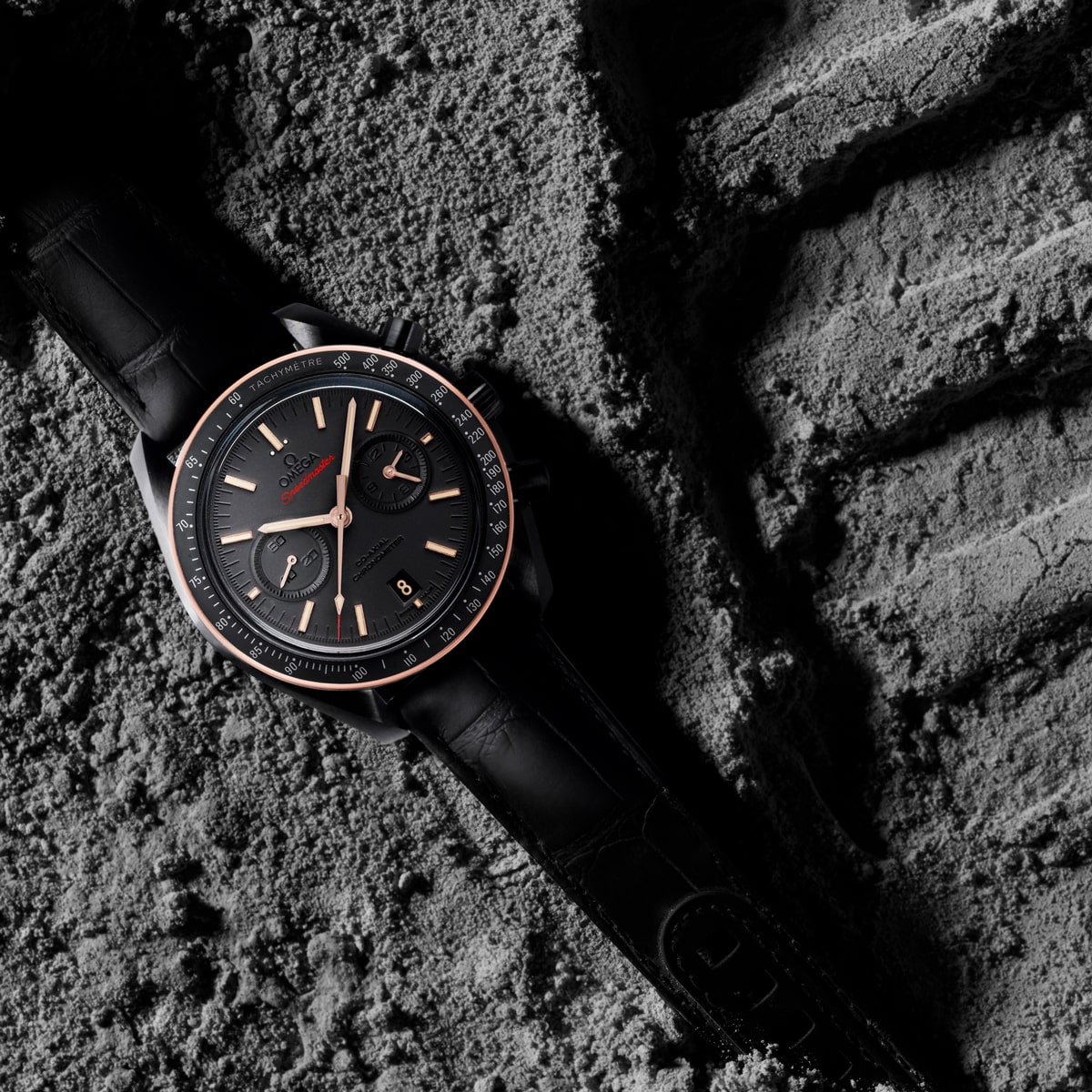 2025 © OMEGA Eine schwarze Armbanduhr mit rundem Zifferblatt und roségoldenen Akzenten liegt auf einem grauen, felsigen Untergrund, der an die geheimnisvolle dunkle Seite des Mondes erinnert. Die Uhr verfügt über ein schwarzes Lederarmband und detaillierte Markierungen auf dem Zifferblatt.