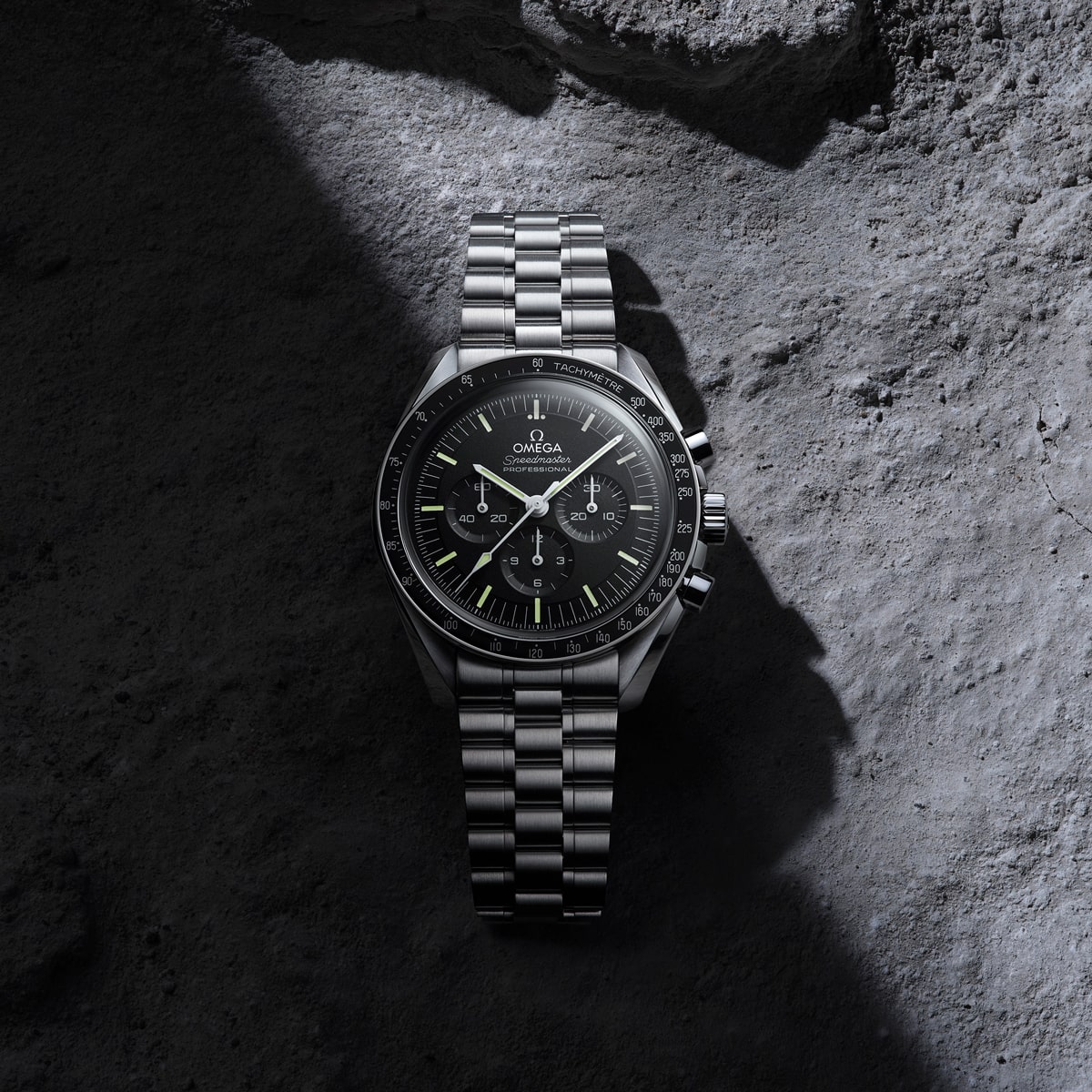 2025 © OMEGA Eine Omega Speedmaster Moonwatch aus Edelstahl mit schwarzem Zifferblatt und Chronographenfunktionen ruht auf einer rauen, grauen Felsoberfläche, die teilweise von einem Lichtschacht beleuchtet wird.
