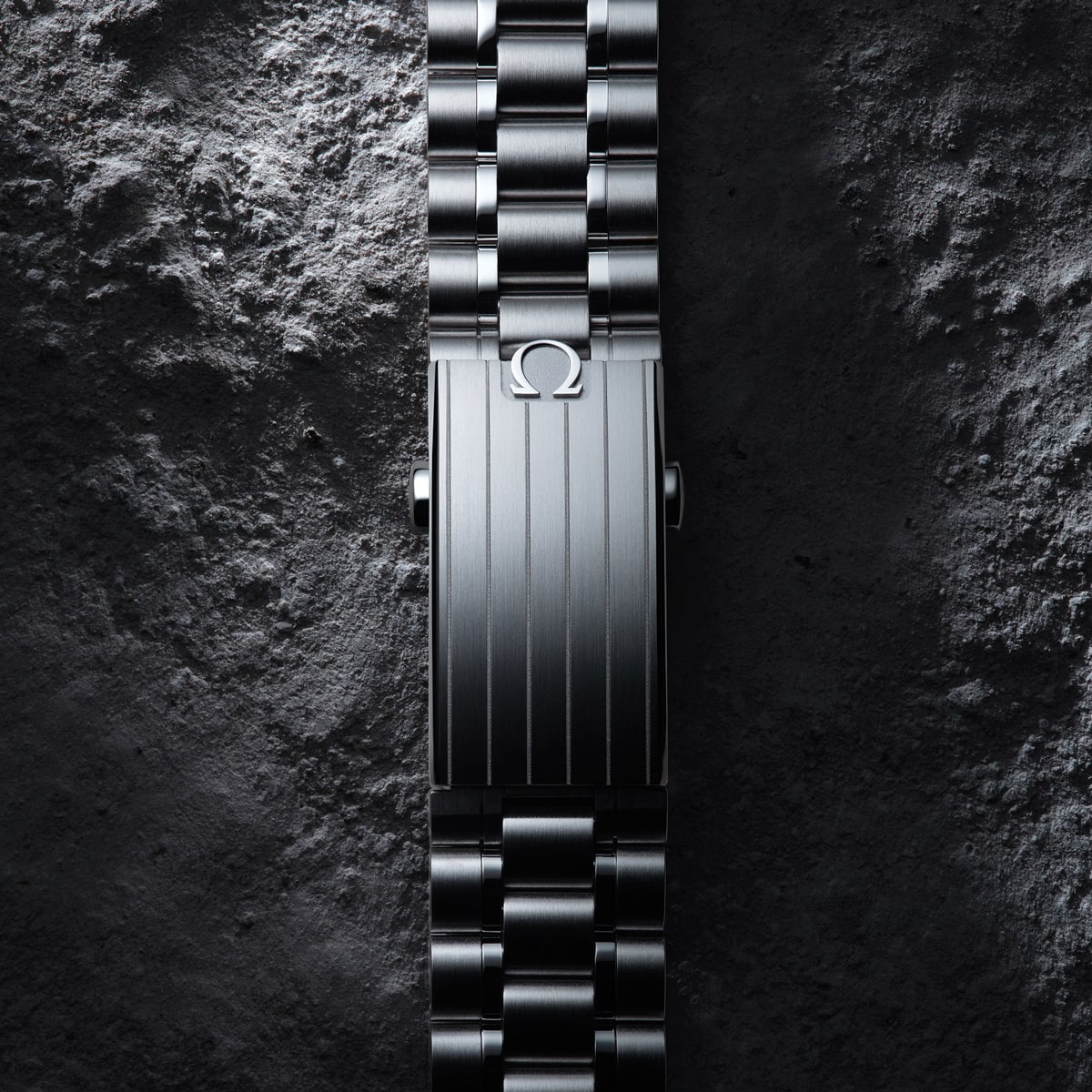 2025 © OMEGA Nahaufnahme einer metallischen Omega Moonwatch-Armbandschließe mit vertikalen Rillen und dem Omega-Logo auf einem strukturierten, dunklen Steinhintergrund.
