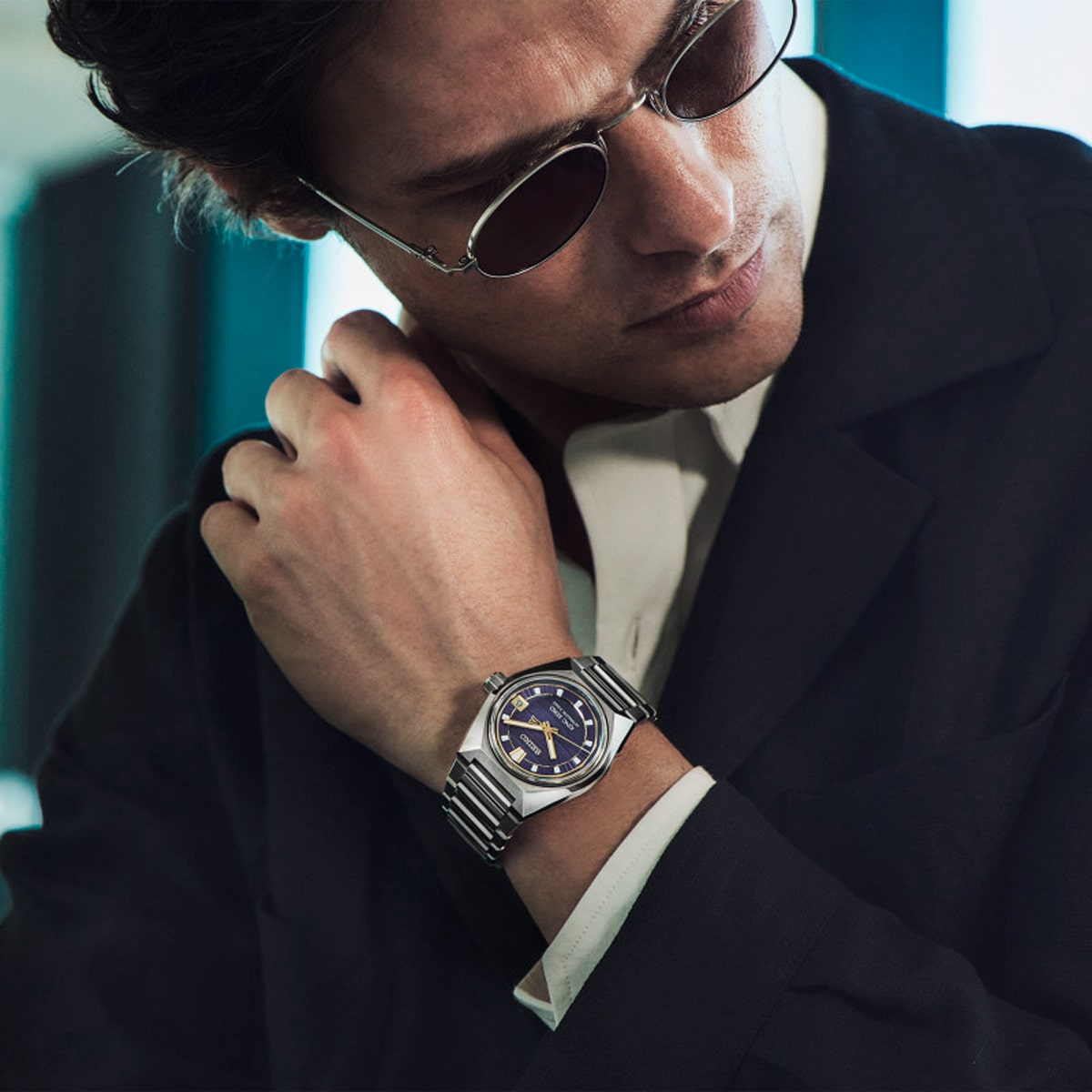 2025 © SEIKO Ein Mann mit Sonnenbrille und dunklem Anzug blickt auf seine silberne Seiko-Armbanduhr mit blauem Zifferblatt und hält mit einer Hand seinen Kragen fest. Der Hintergrund ist sanft verschwommen mit blaugrünen und weißen Tönen.