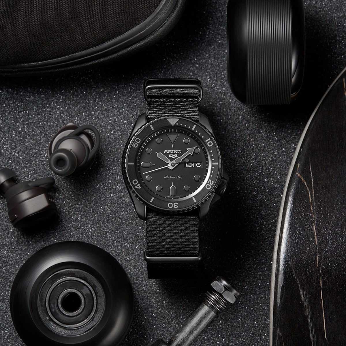2025 © SEIKO Eine elegante Seiko-Armbanduhr mit schwarzem Nylonarmband ruht auf einer dunklen, strukturierten Oberfläche, umgeben von Skateboard-Rädern, einem Skateboard und kabellosen Kopfhörern. Die Seiko-Uhr zeigt die Uhrzeit, den Tag und das Datum an.