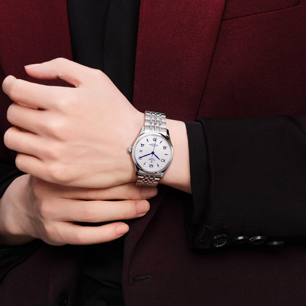 2025 © TUDOR Eine Person, die einen burgunderroten Blazer und ein schwarzes Hemd trägt, zeigt eine silberne Armbanduhr von Tudor 1926 mit weißem Zifferblatt und blauen Zeigern an ihrem linken Handgelenk und hält sie mit der rechten Hand fest.