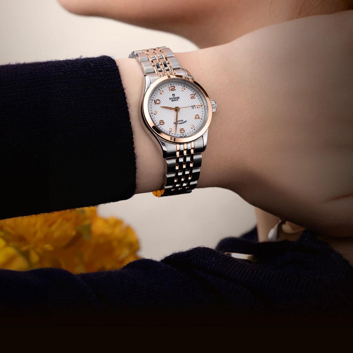 2025 © TUDOR Nahaufnahme des Handgelenks einer Person, die eine silberne und goldene Omega- oder Tudor-Armbanduhr von 1926 mit weißem Zifferblatt und goldenen Indexen trägt. Der Hintergrund ist unscharf, gelbe Blumen sind zu sehen.