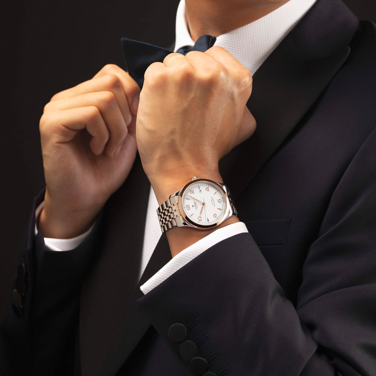 2025 © TUDOR Eine Person in einem dunklen Anzug und weißem Hemd rückt eine schwarze Fliege zurecht. Am linken Handgelenk trägt sie eine silberne Armbanduhr von Tudor 1926 mit weißem Zifferblatt und Metallband. Der Hintergrund ist dunkel.