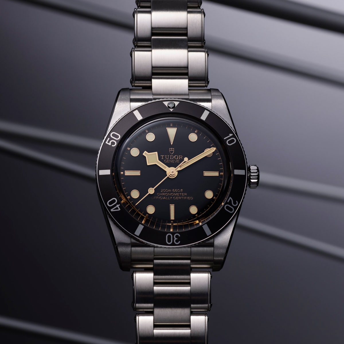 2025 © TUDOR Eine Tudor Black Bay 54 Armbanduhr mit schwarzer Lünette, schwarzem Zifferblatt und silbernem Metallarmband wird vor einem diagonal gestreiften, metallgrauen Hintergrund präsentiert. Die Uhr verfügt über goldene Stundenmarkierungen und Zeiger.