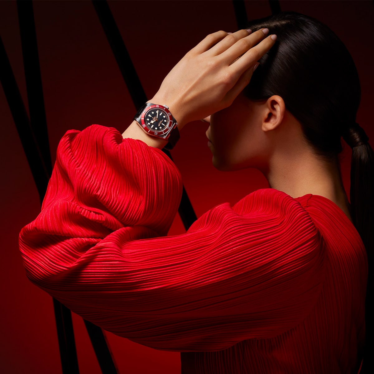 2025 © TUDOR Eine Person in einer strukturierten roten Bluse bedeckt einen Teil ihres Gesichts mit einer Hand und präsentiert eine auffällige Black Bay-Armbanduhr in Schwarz und Rot. Der Hintergrund ist dunkel mit roter Beleuchtung und schwarzen abstrakten Linien.