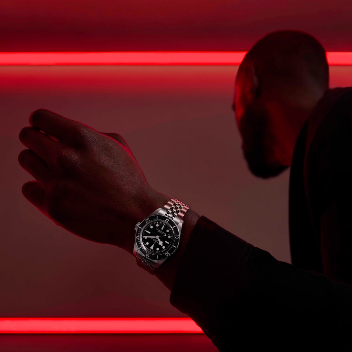 2025 © TUDOR Eine Person mit Bart, die als Silhouette dargestellt ist, streckt ihren Arm aus, um eine silberne Black-Bay-Luxusarmbanduhr zu präsentieren. Rote Neonröhren bilden horizontale Linien im Hintergrund und verleihen der Szene einen dramatischen Effekt.