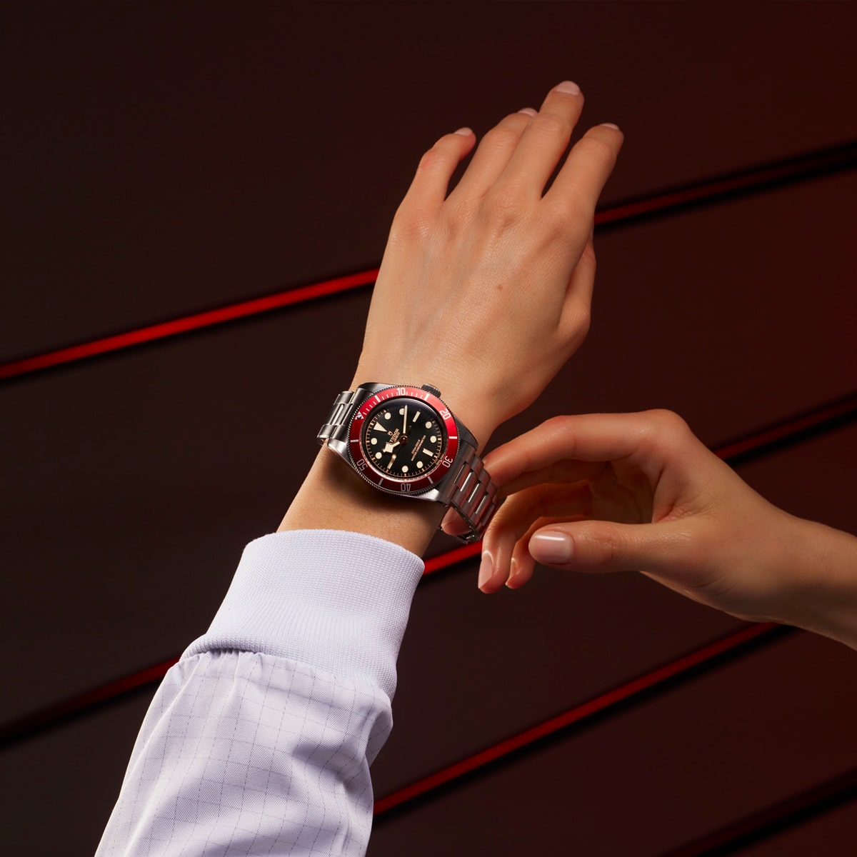 2025 © TUDOR Nahaufnahme einer Person, die eine silberne Black Bay-Armbanduhr mit schwarzer und roter Lünette trägt. Ein anderer Zeiger stellt die Uhr vor einem dunklen Hintergrund mit auffälligen diagonalen roten Streifen ein.