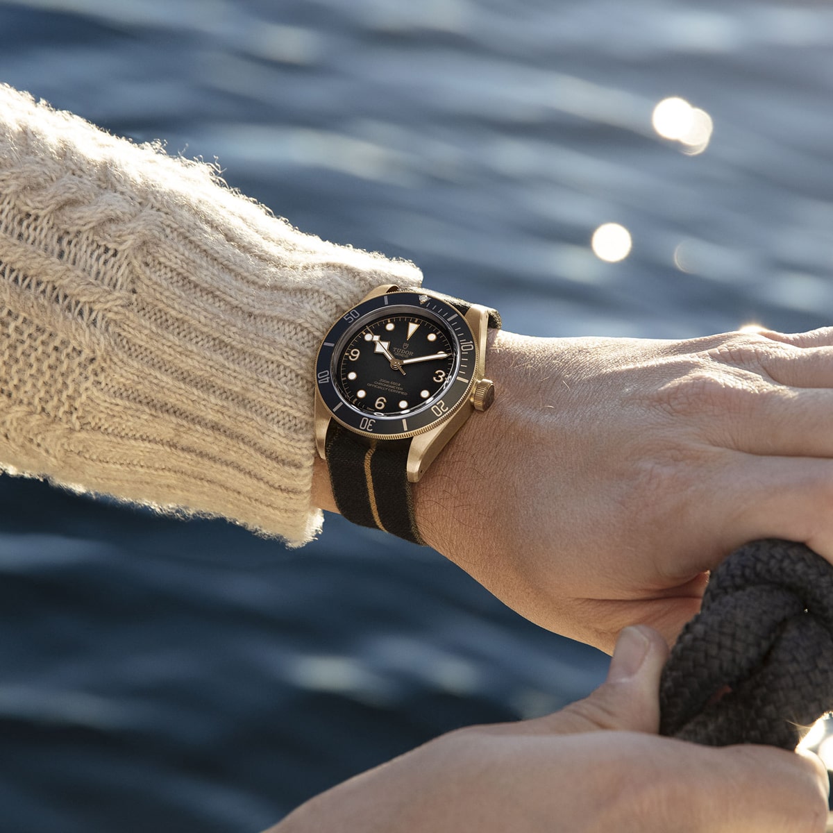 2025 © TUDOR Eine Person in einem beigen Strickpullover hält ein Seil in der Nähe des Wassers und zeigt eine Black Bay Bronze-Armbanduhr mit schwarzem Armband an ihrem linken Handgelenk. Das Sonnenlicht spiegelt sich auf dem plätschernden Wasser im Hintergrund.
