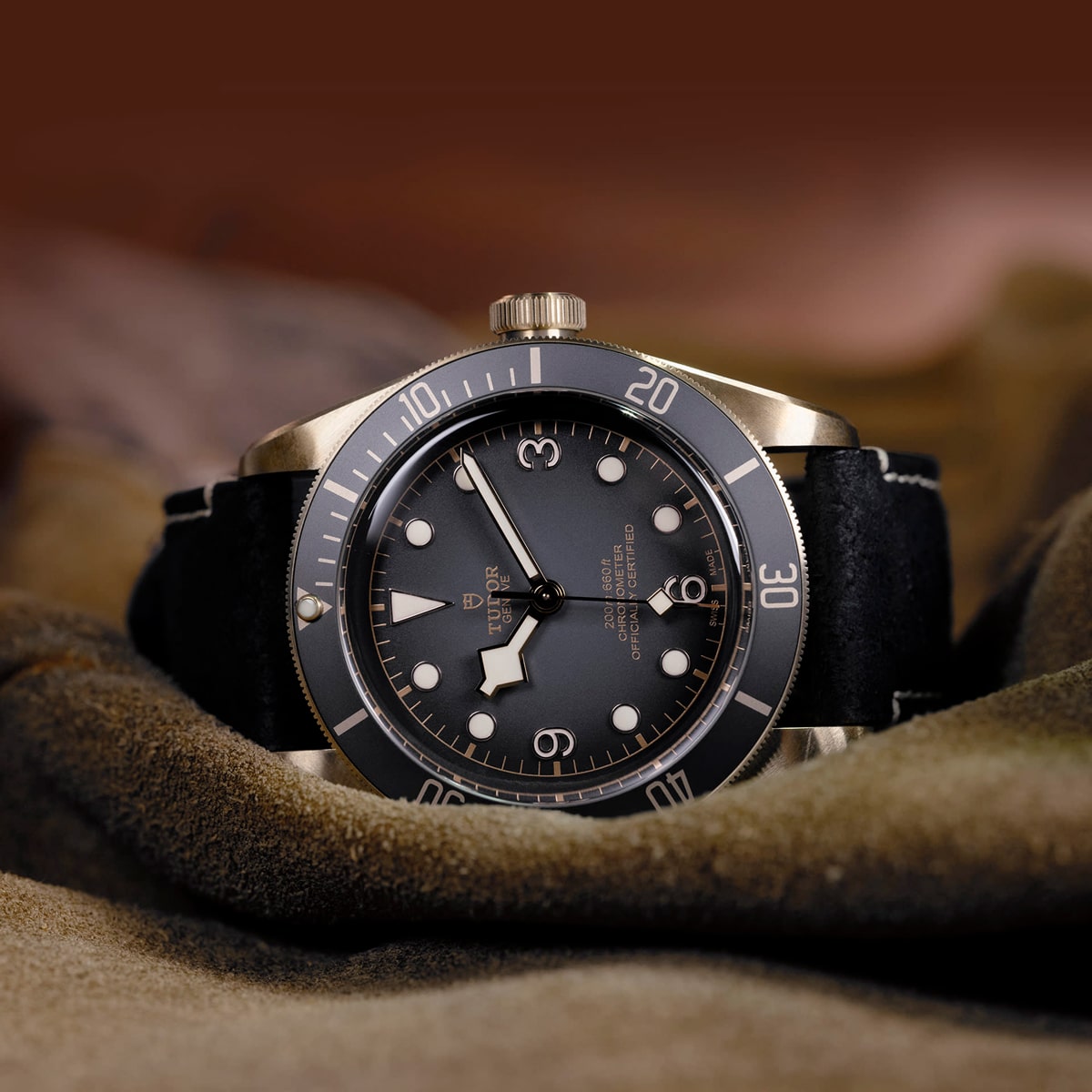 2025 © TUDOR Eine Nahaufnahme einer luxuriösen Black Bay Bronze-Armbanduhr mit schwarzer Lünette und schwarzem Zifferblatt, silbernen Indexen und einem schwarzen Lederarmband, das auf einem weichen braunen Stoff mit einem unscharfen braunen Hintergrund ruht.
