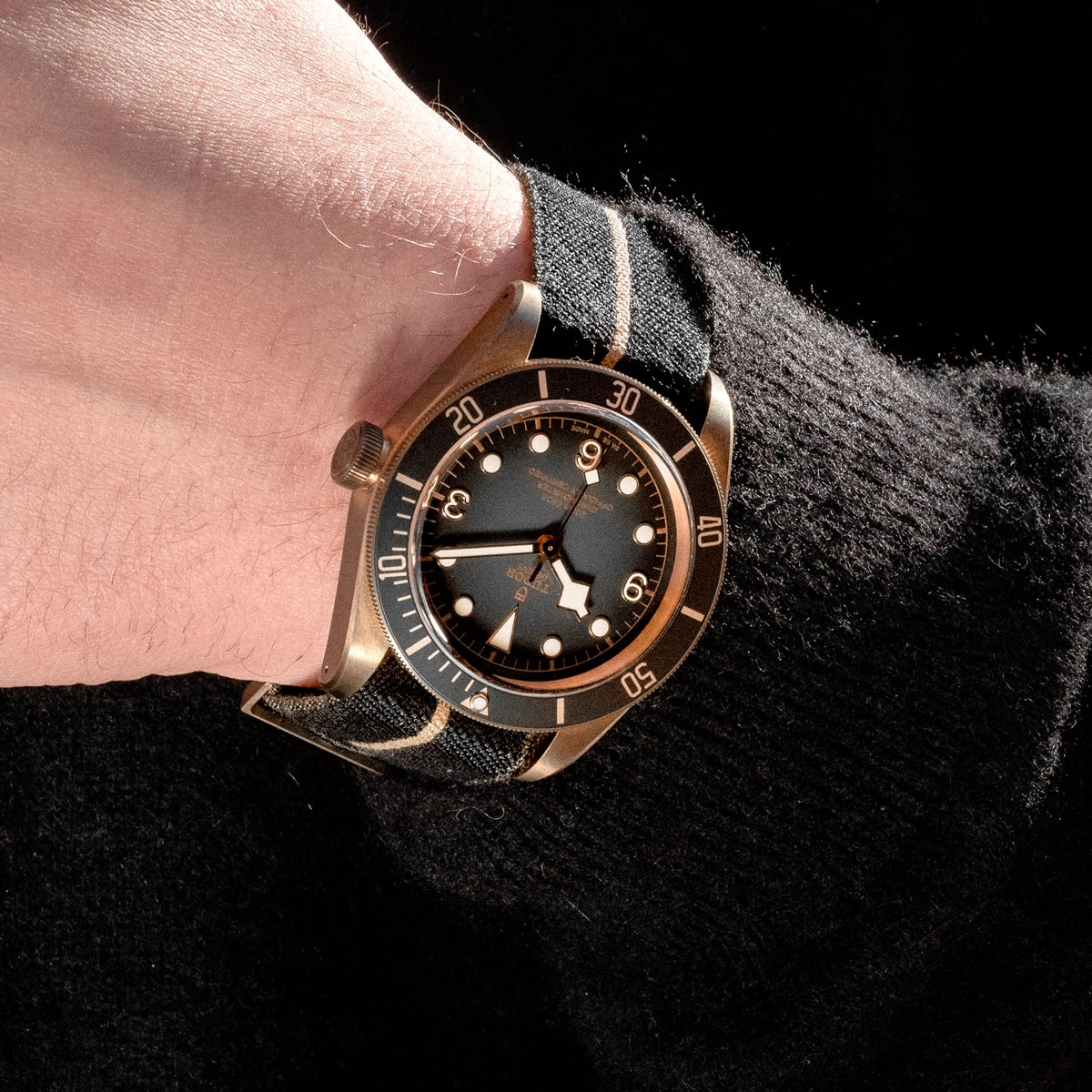 2025 © TUDOR Nahaufnahme eines Handgelenks mit einer Black Bay Bronze-Uhr mit Stoffarmband, schwarzem Zifferblatt, goldenen Stundenmarkierungen und Leuchtzeigern auf einer schwarzen, strukturierten Hülle.