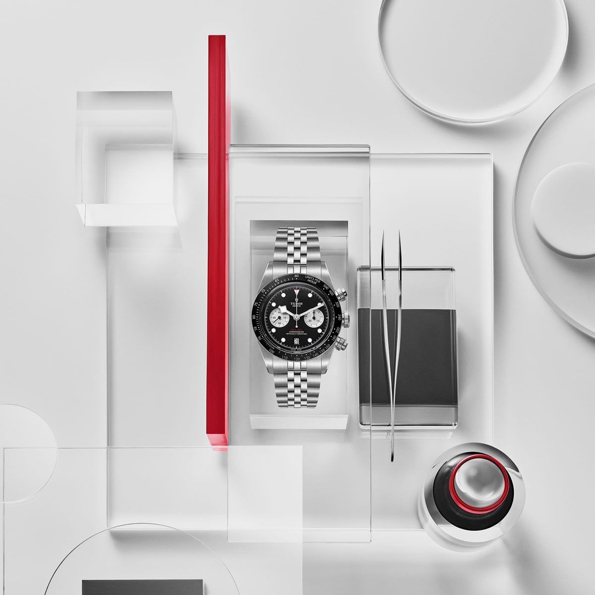 2025 © TUDOR Eine silberne Black Bay Chrono Armbanduhr mit schwarzem Ziffernblatt ist zwischen geometrischen Glasformen, roten Akzenten und minimalistischem Geschirr auf weißem Hintergrund zu sehen. Die Szene ist modern und abstrakt.