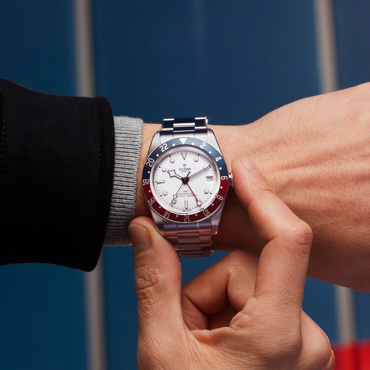 2025 © TUDOR Eine Person, die eine silberne Tudor Black Bay GMT-Armbanduhr mit blauer und roter Lünette, weißem Zifferblatt und Metallarmband trägt, stellt die Uhr an ihrem linken Handgelenk ein. Im Hintergrund sind unscharfe blaue vertikale Flächen zu sehen.