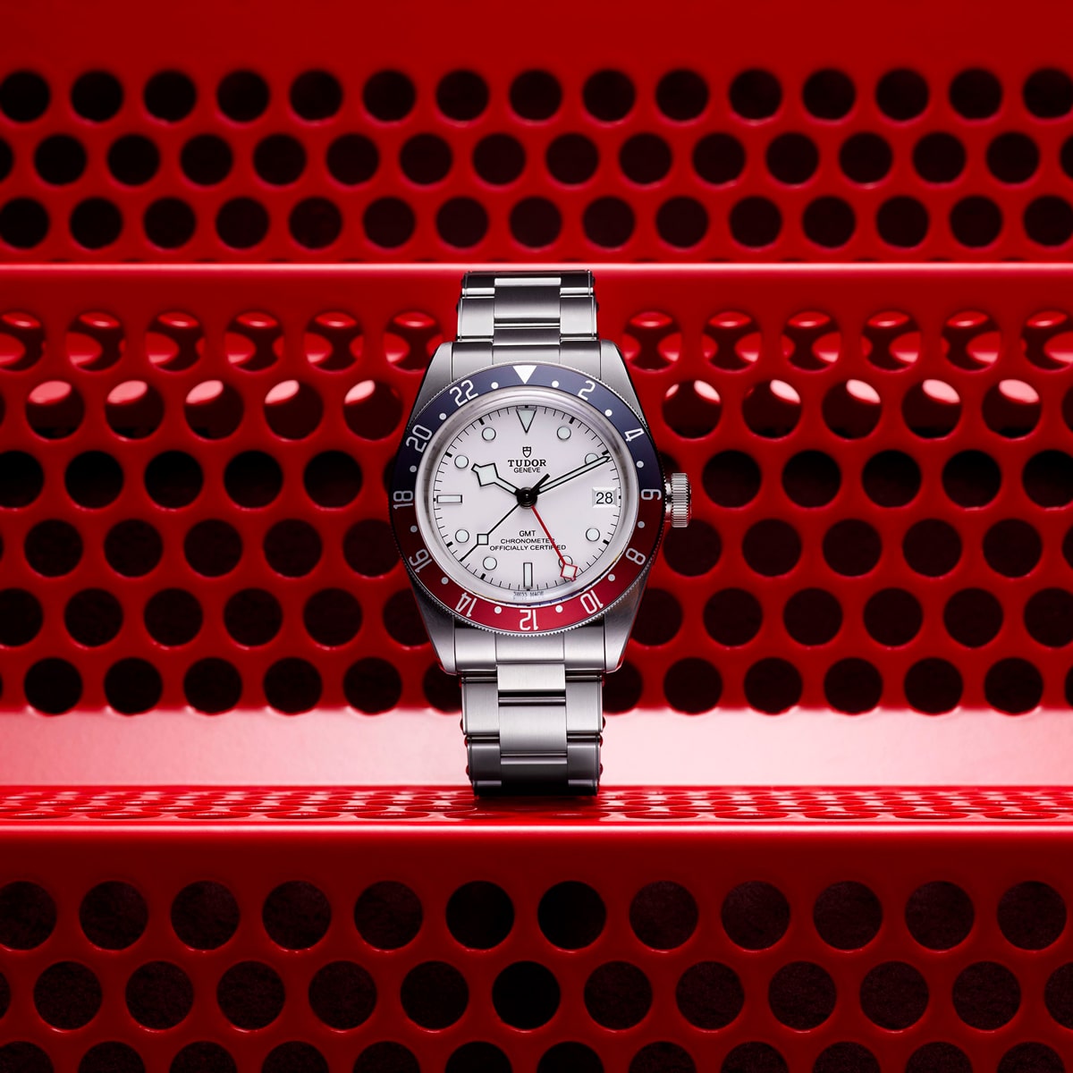 2025 © TUDOR Eine Tudor Black Bay GMT Armbanduhr mit silbernem Metallband und rot-blauer Lünette steht aufrecht auf einer perforierten roten Metallfläche mit kreisförmigen Löchern. Der Hintergrund besteht ebenfalls aus rotem, perforiertem Metall.