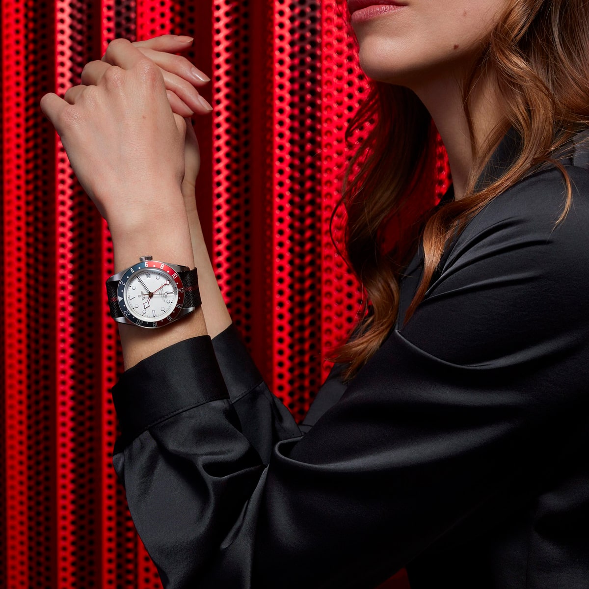 2025 © TUDOR Eine Frau in einem schwarzen Hemd posiert mit verschränkten Armen und präsentiert eine Black Bay GMT-Armbanduhr mit weißem Zifferblatt und roter und blauer Lünette vor einem leuchtend roten, perforierten Hintergrund. Ihr Gesicht ist teilweise sichtbar.