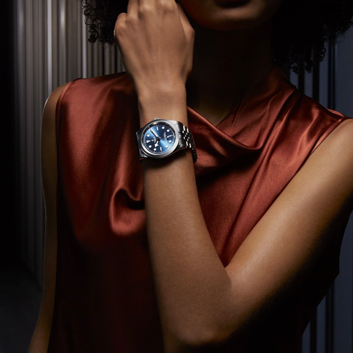 2025 © TUDOR Eine Person, die ein ärmelloses, glänzendes kupferfarbenes Oberteil trägt, zeigt eine von der Black Bay One inspirierte silberne Armbanduhr mit blauem Zifferblatt an ihrem linken Handgelenk. Der Hintergrund ist schwach beleuchtet und weist vertikale metallische Linien auf.