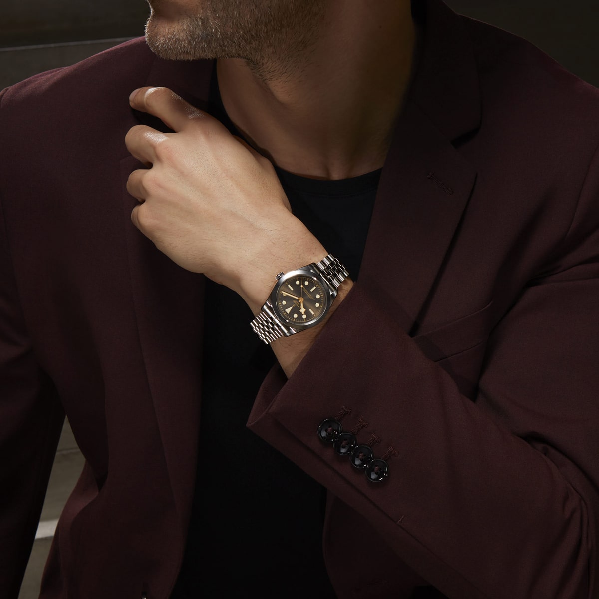 2025 © TUDOR Ein Mann in einem weinroten Anzug und einem schwarzen Hemd hält seine Hand in den Nacken und zeigt eine Black Bay One-Armbanduhr mit Metallarmband und dunklem Zifferblatt. Sein Gesicht ist teilweise sichtbar.