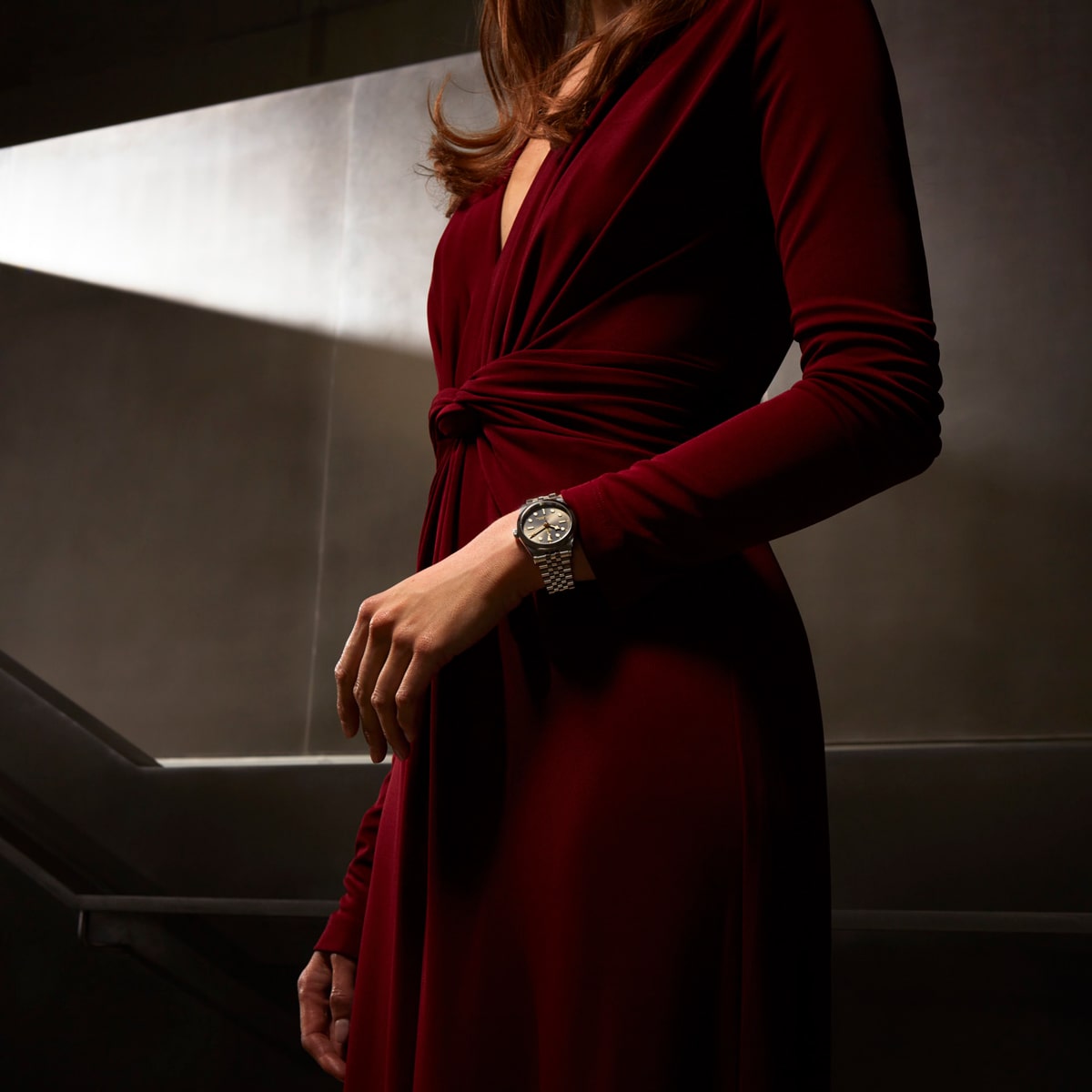 2025 © TUDOR Eine Frau in einem langärmeligen, tiefroten Kleid steht in einem Innenraum, ihr Gesicht ist nicht sichtbar. Sie trägt eine von der Black Bay One inspirierte silberne Armbanduhr mit rundem Ziffernblatt, die in einer modernen, schwach beleuchteten Umgebung deutlich an ihrem linken Handgelenk zu erkennen ist.