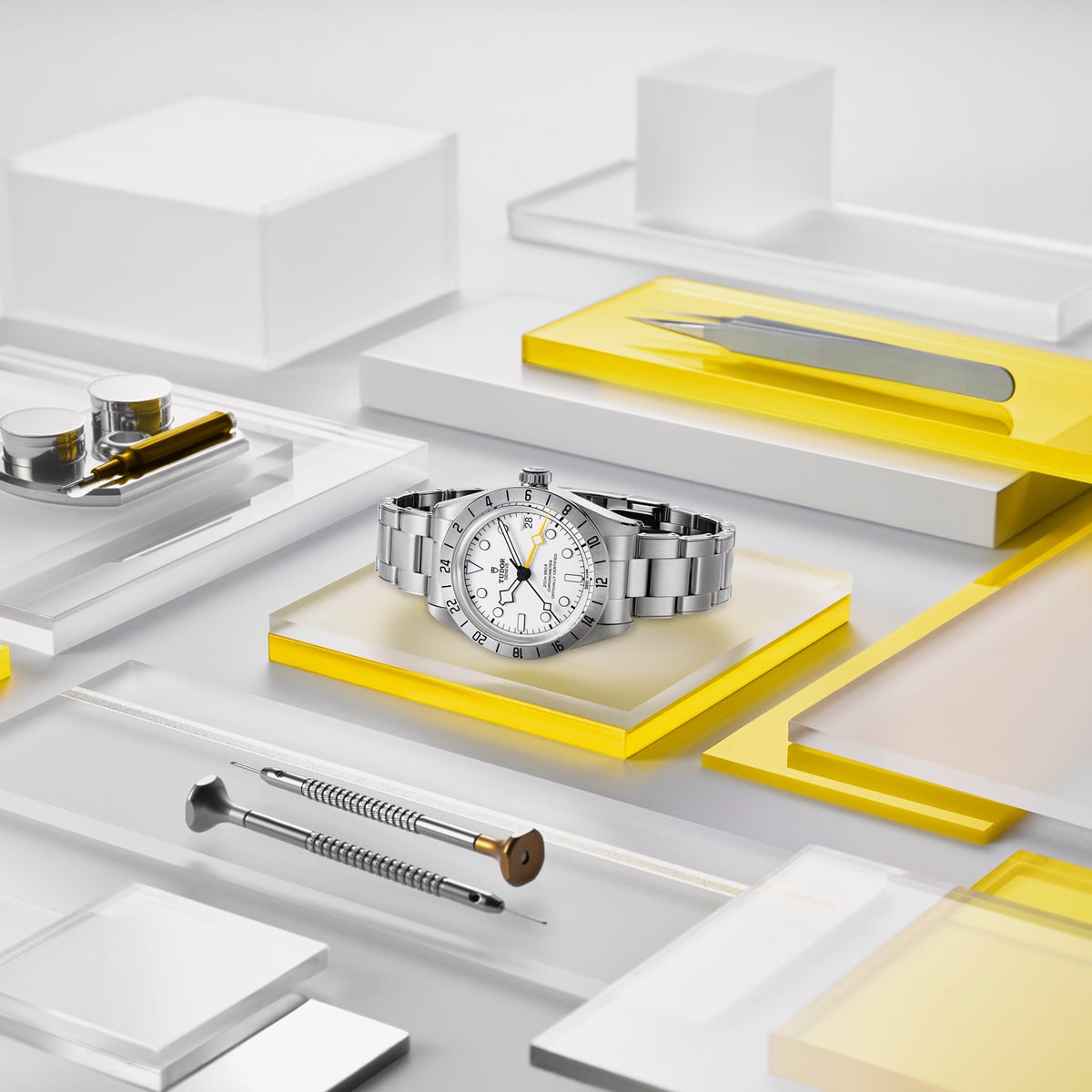 2025 © TUDOR Eine silberne Black Bay Pro-Armbanduhr mit weißem Zifferblatt steht auf einem gelben und durchsichtigen Podest, umgeben von Uhrmacherwerkzeugen und -behältern, die auf geometrischen, transparenten und gelben Flächen übersichtlich angeordnet sind.