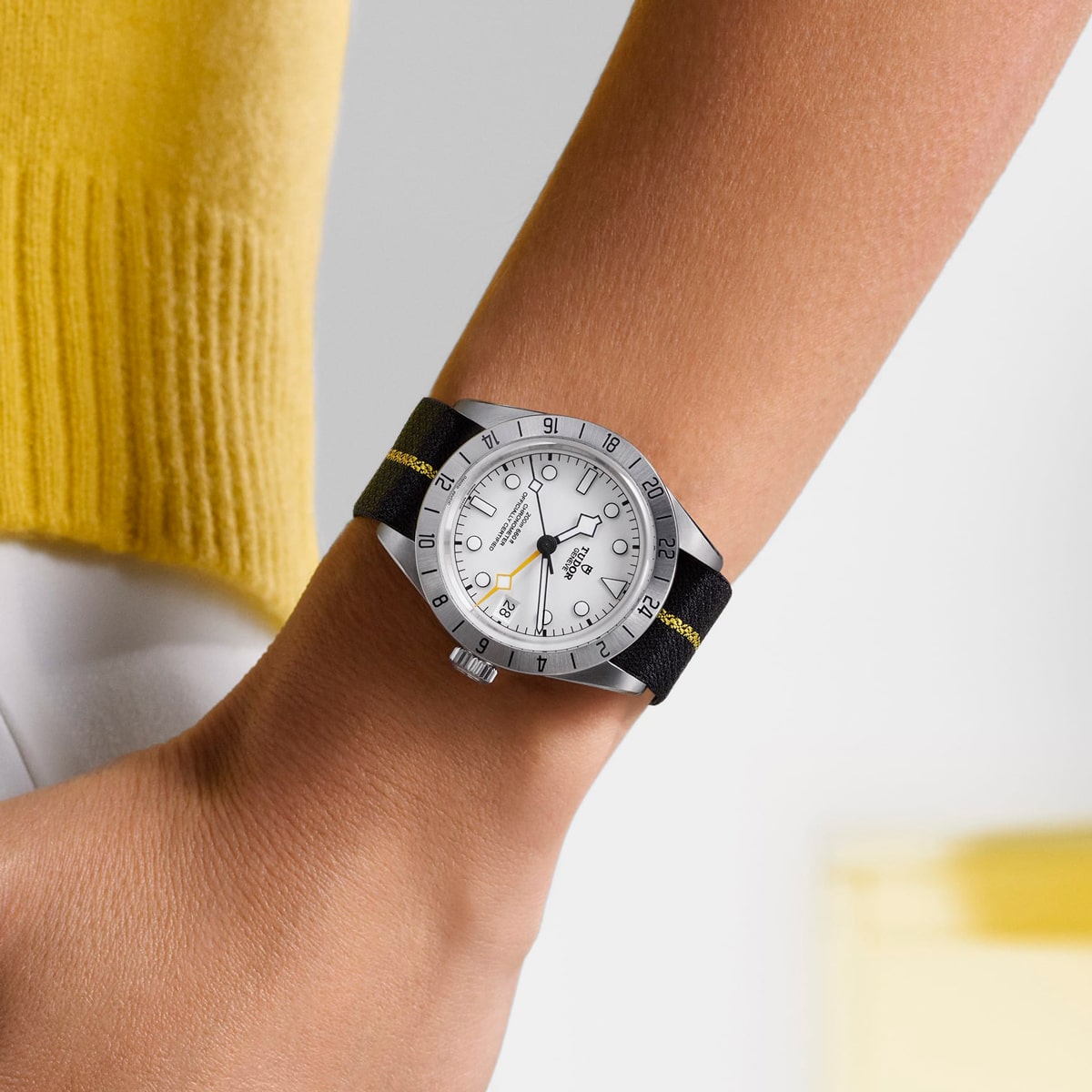 2025 © TUDOR Nahaufnahme des Handgelenks einer Person, die eine silberne Black Bay Pro-Armbanduhr mit weißem Zifferblatt und schwarzem Armband trägt. Die Person ist mit einem gelben Oberteil und einer weißen Hose bekleidet und steht vor einem minimalen, hellen Hintergrund.