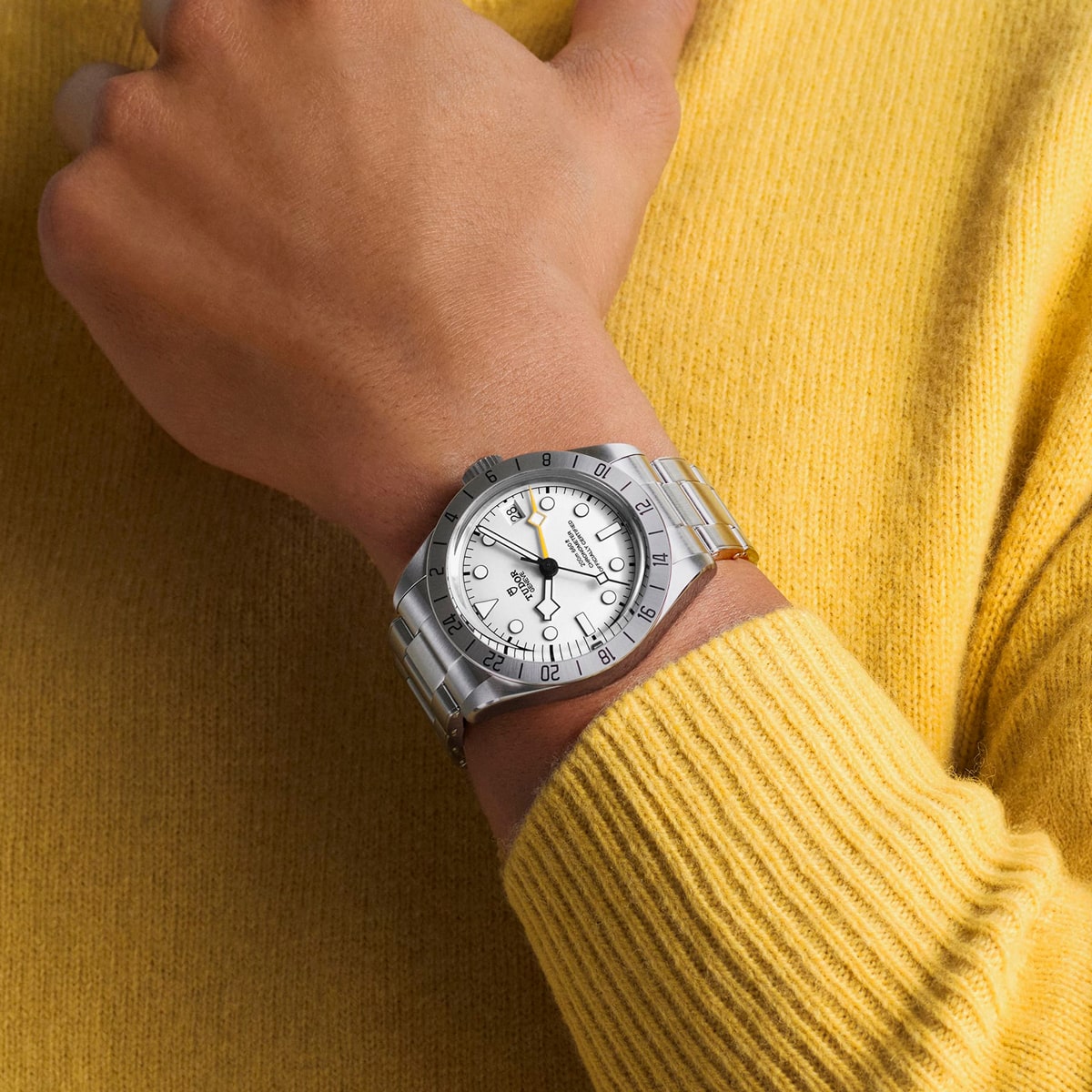 2025 © TUDOR Eine Person, die einen gelben Pullover trägt, zeigt eine silberne Black Bay Pro-Armbanduhr mit weißem Zifferblatt, schwarzen Stundenmarkierungen und einem Metallgliederband an ihrem linken Handgelenk.