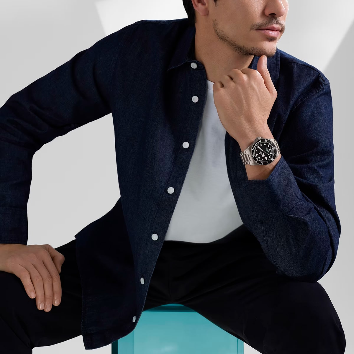 Juwelier-Wenthe-Uhren-Schmuck-Mannheim-TUDOR-Pelagos-Ansicht-1 Ein Mann, der ein dunkles Jeanshemd über einem weißen T-Shirt und eine schwarze Hose trägt, sitzt auf einem durchsichtigen blauen Hocker und zeigt eine silberne Pelagos-Armbanduhr mit schwarzem Zifferblatt an seinem linken Handgelenk. Seine rechte Hand ruht nachdenklich auf seinem Kinn.