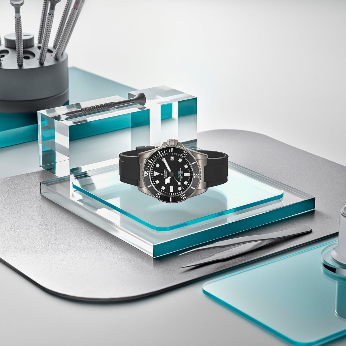 2025 © TUDOR Eine schwarze Pelagos-Armbanduhr mit silberner Lünette wird auf einer transparenten Glasplattform präsentiert, umgeben von Uhrmacherwerkzeugen aus Metall und weiteren Glasblöcken auf einer schlichten, modernen Arbeitsfläche.