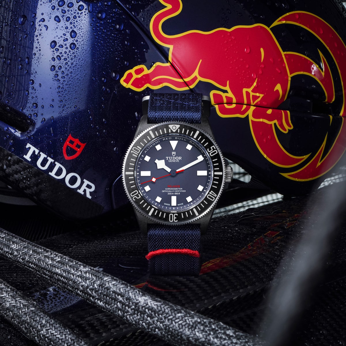 2025 © TUDOR Eine Tudor Pelagos FXD Armbanduhr mit marineblauem Armband ist vor einem Rennhelm platziert, der ein rotes und gelbes Stierlogo, Wassertropfen und das Tudor-Logo auf einer strukturierten Oberfläche zeigt.