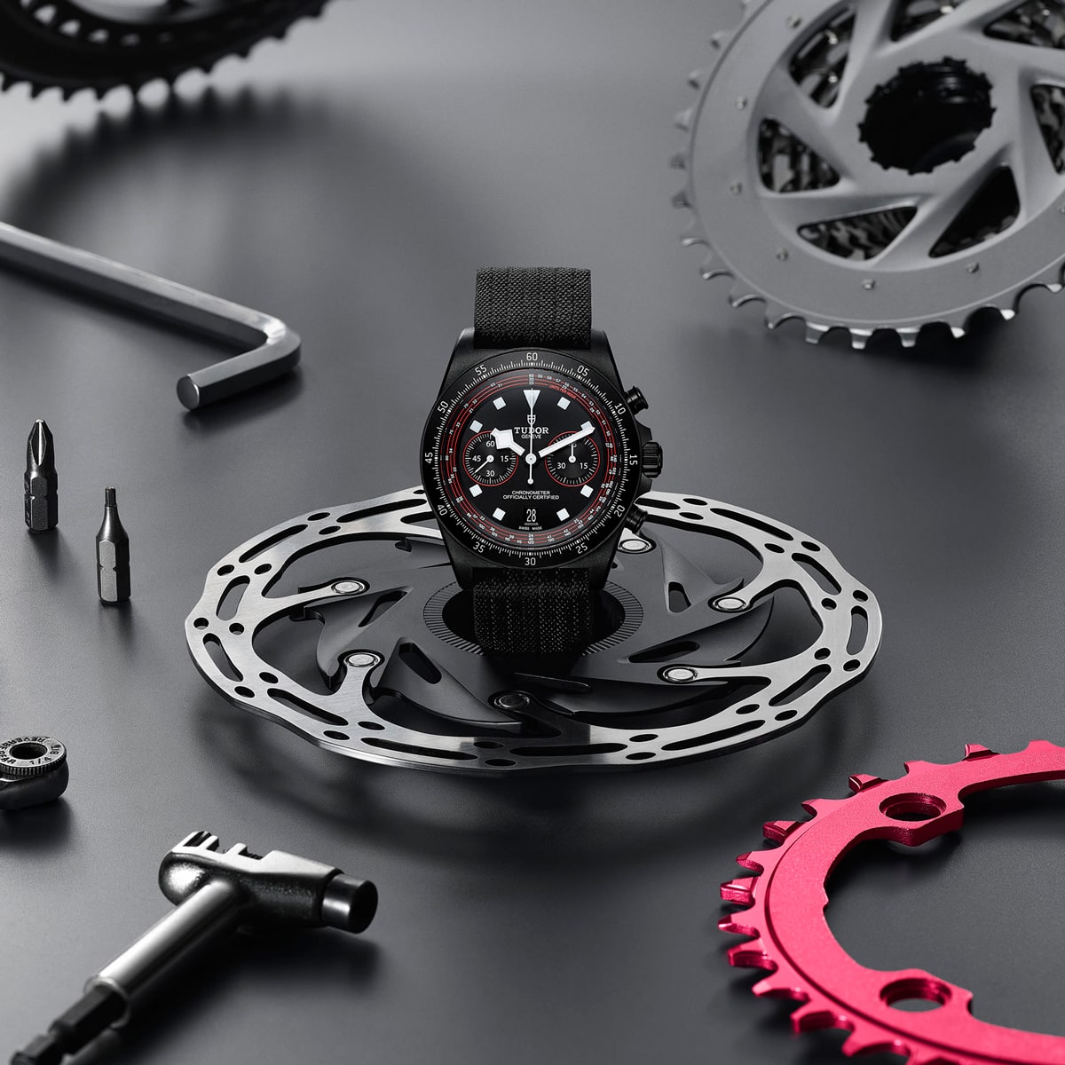 2025 © TUDOR Eine schwarze Pelagos FXD-Armbanduhr mit Stoffarmband ist auf einem metallischen Fahrradscheibenbremsrotor abgebildet, umgeben von Fahrradschaltungen, Sechskantwerkzeugen, Schraubendreherbits und einem roten Zahnrad auf grauem Hintergrund.