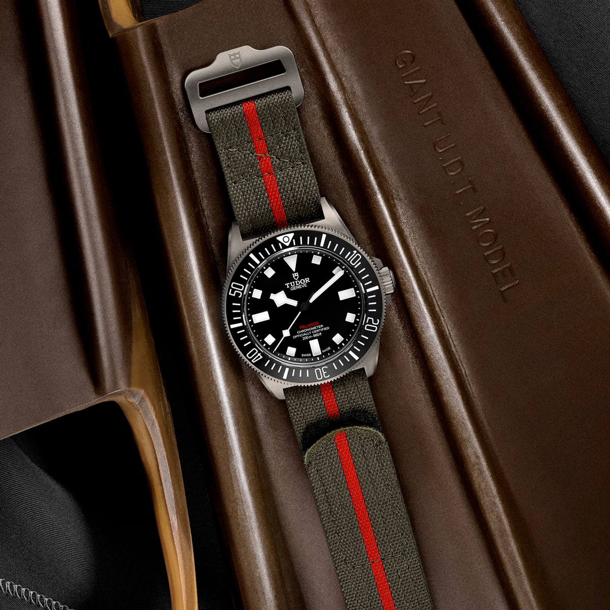 2025 © TUDOR Eine Tudor Pelagos FXD Taucheruhr mit schwarzem Zifferblatt und markanten weißen Markierungen auf einem olivgrünen Stoffarmband mit rotem Streifen, auf einem braunen Gehäuse mit der Aufschrift "GIANT U.D.T. MODEL.