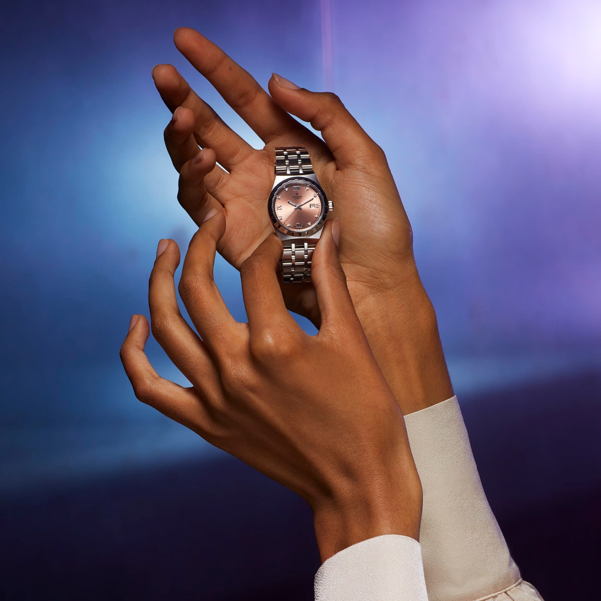 2025 © TUDOR Zwei Hände halten zart eine silberne Tudor Royal Armbanduhr mit roségoldenem Zifferblatt vor einem verschwommenen blauen und violetten Hintergrund. Die Person trägt ein weißes, langärmeliges Kleidungsstück.