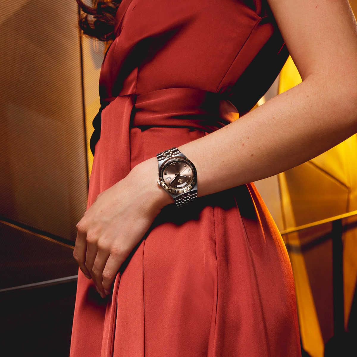 2025 © TUDOR Eine Frau in einem roten Kleid steht mit angewinkeltem Arm und präsentiert eine silberne Tudor Royal-Armbanduhr. Warmes, goldenes Licht unterstreicht die elegante und stilvolle Szene.