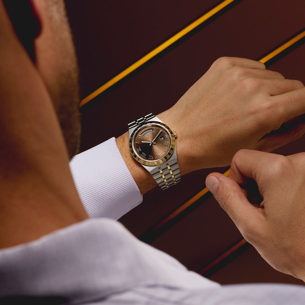 2025 © TUDOR Eine Person mit weißem Hemd prüft die Uhrzeit auf einer zweifarbigen Luxus-Armbanduhr von Tudor Royal mit goldenem und silbernem Armband vor einem braunen Hintergrund mit diagonalen gelben Linien.