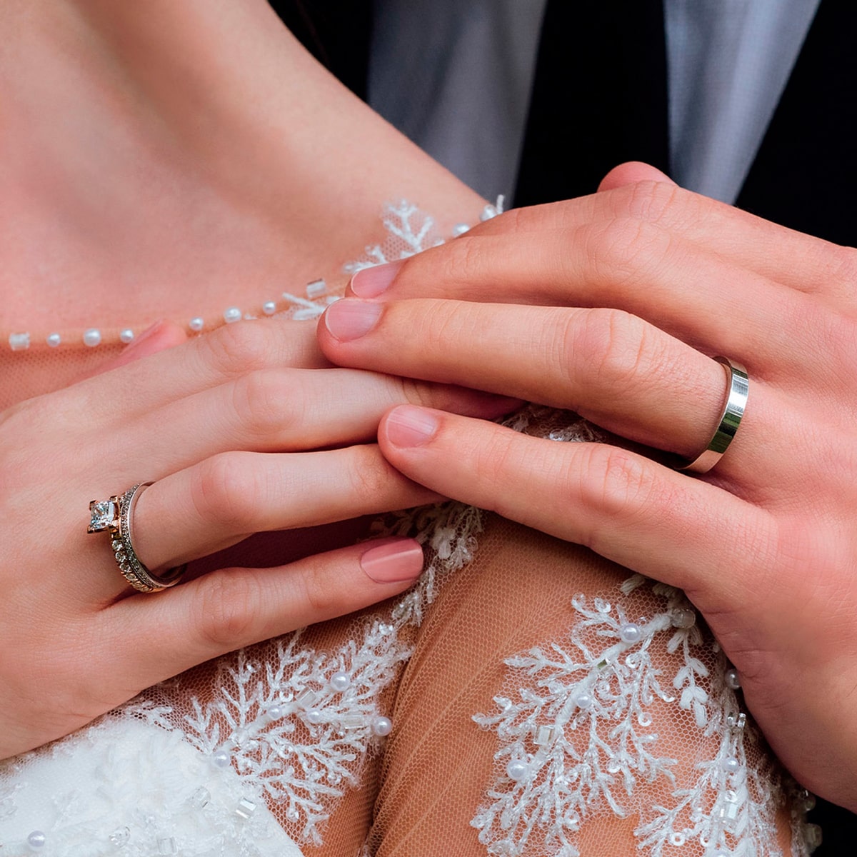 2025 © 123GOLD Nahaufnahme zweier Hände mit 123GOLD Trauringe, die sich sanft berühren. Sie tragen elegante Kleidung mit zarter Spitze und Perlendetails, die auf eine Hochzeit oder einen besonderen Anlass hinweisen.