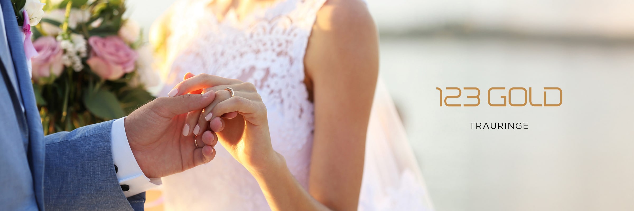 2025 © 123GOLD Eine Braut und ein Bräutigam tauschen im Freien die Ringe aus; die Braut trägt ein weißes Spitzenkleid und hält einen Blumenstrauß. Neben den beiden steht "123GOLD Trauringe" auf einem unscharfen hellen Hintergrund.