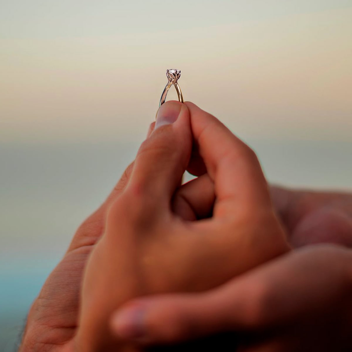 2025 © 123GOLD Eine Nahaufnahme von Händen, die einen 123GOLD Verlobungsring mit Diamanten halten, vor einem sanft verschwommenen Hintergrund, der einen romantischen Moment oder einen Heiratsantrag andeutet.