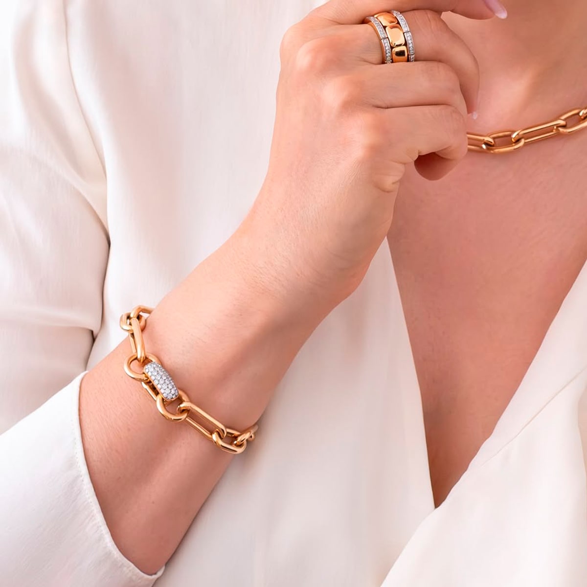 2025 © Al Coro Eine Person, die eine weiße Bluse trägt, zeigt Al Coro Armschmuck - ein goldenes Kettenarmband - zusammen mit einer passenden goldenen Halskette und goldenen Ringen mit eingelassenen Steinen an ihrem Ring- und Mittelfinger.