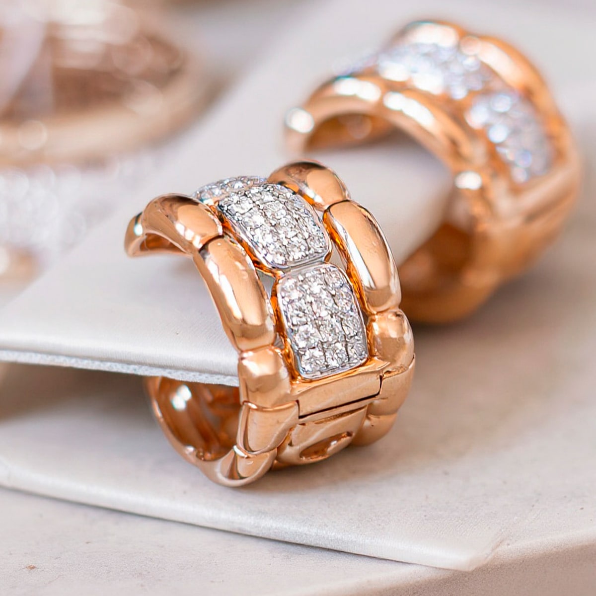 2025 © Al Coro Nahaufnahme von zwei breiten Ringen aus Roségold mit rechteckigem Querschnitt, die mit funkelnden weißen Edelsteinen besetzt sind und auf einer weißen Oberfläche präsentiert werden - eine perfekte Ergänzung zu den Al Coro Ohrringen für einen eleganten, koordinierten Look.