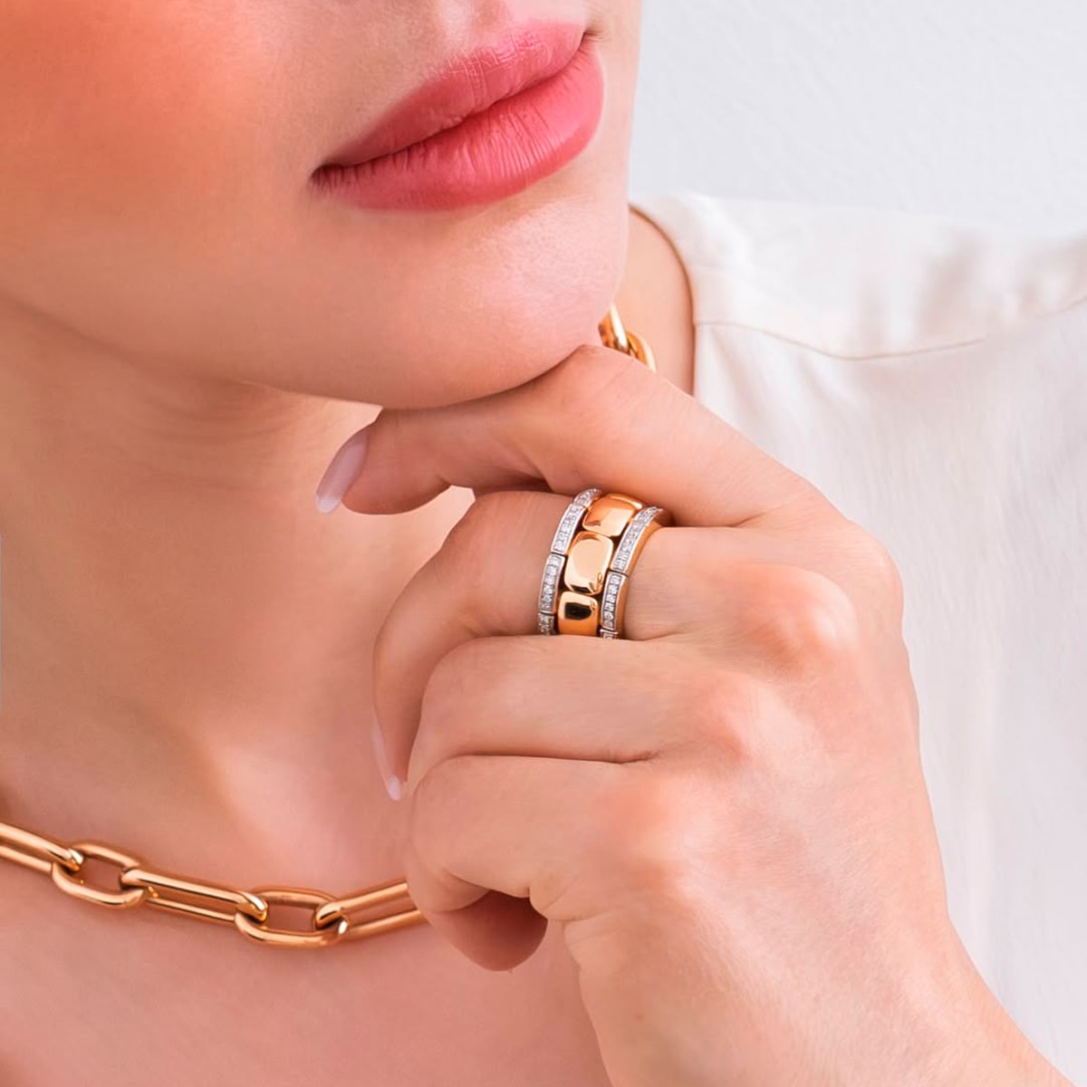 2025 © Al Coro Eine Frau mit rosafarbenen Lippen trägt eine goldene Halskette und zwei gestapelte Al Coro Ringe - Gold- und Diamantringe - an ihrem Finger und stützt ihr Kinn auf ihre Hand. Sie ist mit einem weißen Oberteil bekleidet.